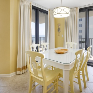 Dining area features a glass-top table with yellow chairs and offers a view of the beach through large sliding doors