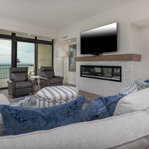 The living area features cozy seating with blue accents, a sleek TV with a fireplace media wall, and stunning beach views