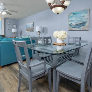 The dining area features a glass table surrounded by gray chairs, with a cozy living space and beach views beyond