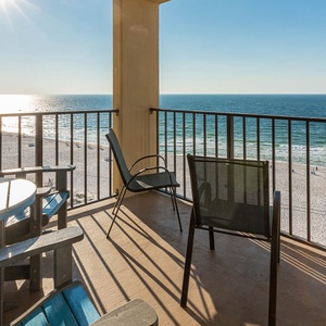 A sunlit balcony features blue Adirondack chairs and a table, offering a serene view of the tranquil beachfront below