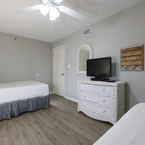This guest bedroom features two double beds, a wicker dresser, and a TV, with calming gray walls and wood flooring