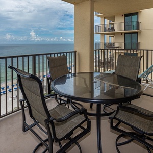 A balcony features a round table with four chairs, offering a serene view of the beach and vibrant blue umbrellas below