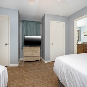 Bright guest bedroom featuring two full beds, a TV on a wicker dresser, and a doorway leading to a well-appointed bathroom