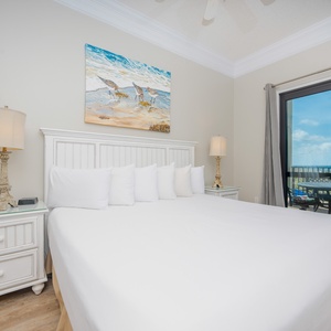 Bright master suite featuring a large king bed with white linens, a lamp, and a balcony offering views of the gulf