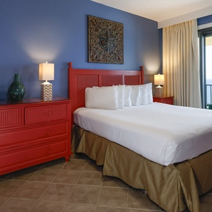 The master bedroom features a red dresser and headboard, with a lovely view of the beach through the balcony doors