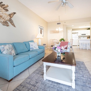 Bright living area featuring a blue sofa with queen sleeper, a wooden coffee table, and a dining space leading to the kitchen
