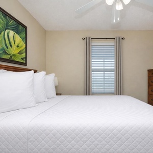 This bedroom features a plush white bed, a wooden dresser, wall-mounted TV, tropical artwork, and natural light