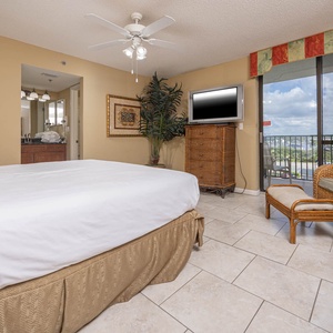 Cozy master bedroom featuring a king bed, a TV, and a seating area with a view of the balcony and lush greenery outside