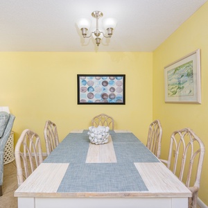 A bright dining area features a large white table with blue accents, surrounded by wicker chairs