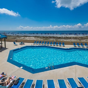 The pool area features a vibrant blue pool surrounded by lounge chairs, with a beach view and a shaded gazebo for relaxation