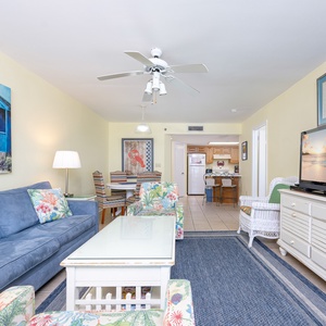 Living area featuring a blue sofa with queen sleeper, colorful chairs, and large TV, leading to a kitchen in the background