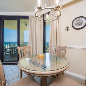 A round glass-top dining table with wooden chairs offers a view of the beach through large sliding glass doors