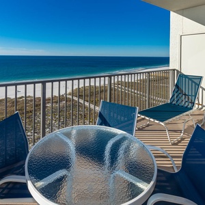 Balcony features a glass-top table and blue chairs, offering a serene view of the gulf and white sand beach below