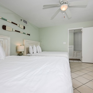 This guest bedroom features two queen beds with white linens, a ceiling fan, and decorative oars on the green wall
