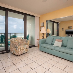 This living area features a turquoise sofa, a patterned chair, and a TV, with a view of the beachfront balcony