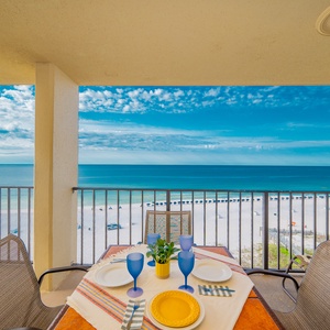 Patio table set for four overlooks the beach front, framed by a vibrant blue sky and calm waters in the distance