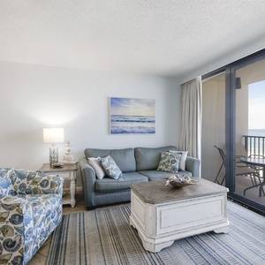 The living area features a comfortable blue sofa, decorative coffee table, and stunning beach view through large glass doors