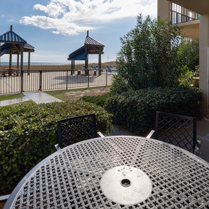 A patio area features a round table surrounded by chairs, with a view of the beach and access to the pool area