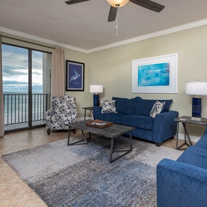 Cozy living area features blue sofas, a patterned chair, and a coffee table, with expansive views of the beach