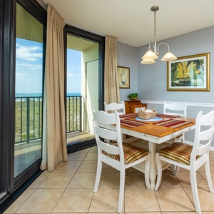 Bright dining area features a wooden table with colorful placemats, surrounded by white chairs, with a view of the beach