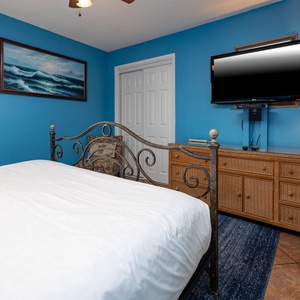 Bright blue walls frame the guest bedroom featuring a king bed, a flat-screen TV, and a stylish wicker dresser