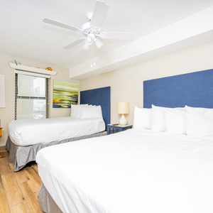 This guest room features two queen beds with blue upholstered headboards, a colorful chair, and a mounted TV
