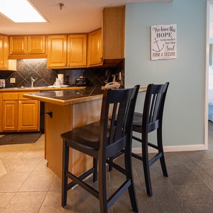 The kitchen features warm wooden cabinetry, a black stove, Keurig, coffee maker, and two high-backed chairs at the bar