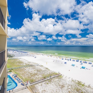 A view from the balcony showcases the vibrant beach with colorful umbrellas and a sparkling pool below, framed by blue skies