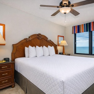 This bedroom features a king bed with plush pillows, a colorful window treatment, and a stunning beachfront view