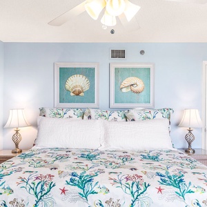 Master bedroom features a colorful king bedt, bedside lamps, and coastal artwork on soft blue walls