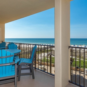 The balcony features vibrant blue chairs and a table, offering a clear view of the beach and calm, turquoise waters