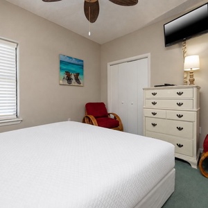 Bright guest bedroom featuring a queen bed, a dresser, a cozy red chair, and a wall-mounted TV for entertainment