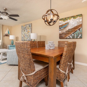 The dining area features a wooden table surrounded by woven chairs, complemented by coastal artwork and a beach view