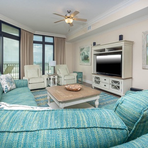 This living area features a cozy turquoise sofa, a large TV, and stunning beachfront views through expansive windows