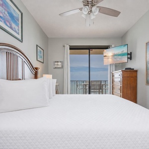 A cozy master bedroom features a wooden headboard, a TV mounted on the wall, and a balcony offering a serene beach view