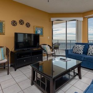 The living room features a blue sofa with patterned cushions and queen sleeper, a flat-screen TV, and a view of the beach
