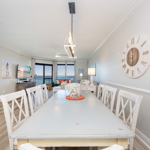 Bright dining area features a large table with white chairs, leading to a cozy living space with a beach view and TV