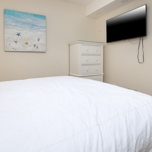 This bedroom features a white quilted bedspread, a mounted TV, and beach-themed artwork for a relaxing atmosphere