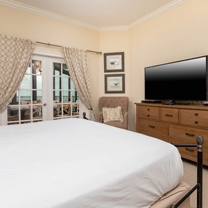 The master bedroom features a cozy king bed with a large TV, inviting armchair, and lovely views through the French doors