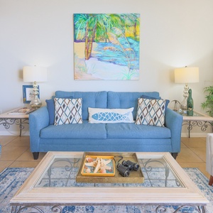 Living area features a blue sofa, a glass coffee table displaying seashells, and vibrant artwork