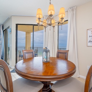 A round wooden dining table with cushioned chairs is illuminated by a stylish chandelier, offering a view of the beach