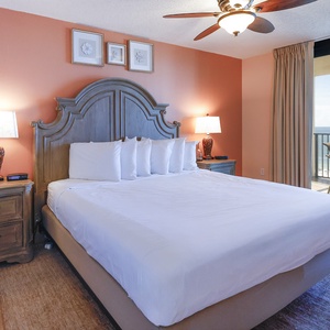 Master bedroom features a king bed with plush pillows, warm orange walls, and a balcony with gulf views