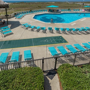 Vibrant turquoise lounge chairs surround a spacious pool area, complete with a shuffleboard court and scenic beach access