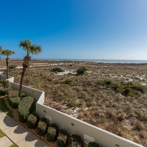 View of the beach with white sand, lush greenery, and a pool area featuring lounge chairs and umbrellas in the distance