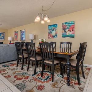 A dining area features a large wooden table surrounded by black chairs, with colorful artwork and a cozy living space in view