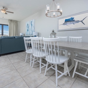 Spacious dining area features a large wooden table with white chairs, complemented by coastal artwork and natural light