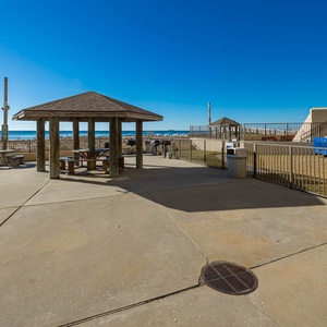 The outdoor pool area features a shaded pavilion with picnic tables, overlooking a beautiful beach front and clear blue skies