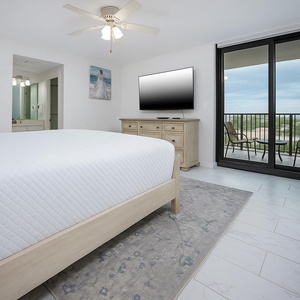Bright master suite featuring a plush king bed, a large TV, and a sliding door leading to a balcony with views of the area