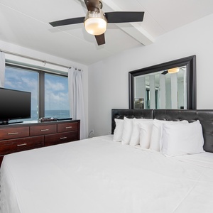 This inviting bedroom features a plush bed, a large TV, and stunning views of the beachfront through expansive windows