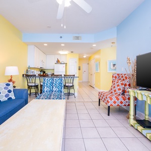 The vibrant living space features a blue sofa, accent chairs, and a large TV, perfect for relaxing after a day at the beach